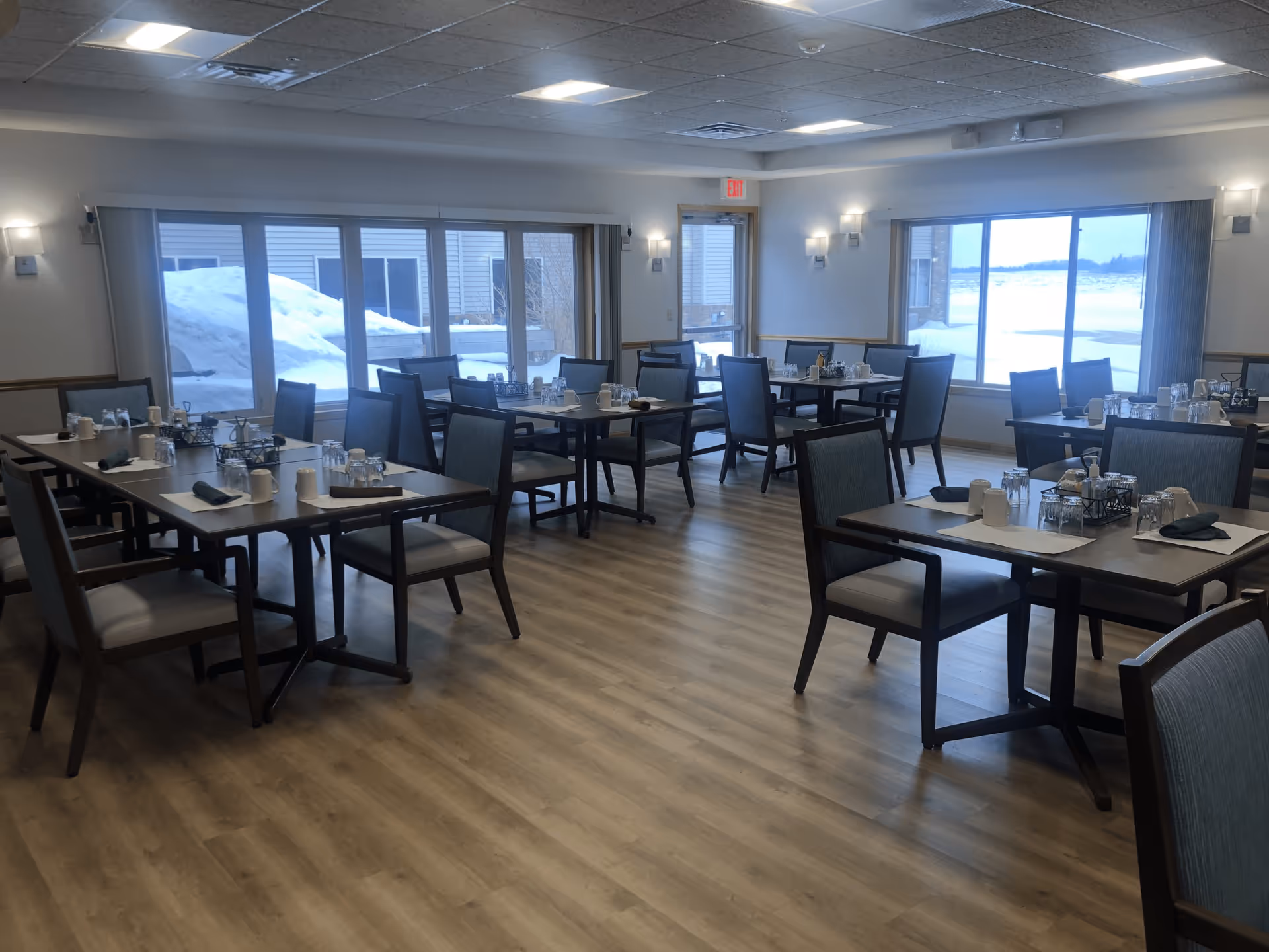 A dining room in a senior living facility with multiple tables and chairs arranged neatly. Each table is set with cups, glasses, napkins, and condiments. Large windows and a glass door provide natural light and a view of snow outside. The room has wooden flooring and soft wall lighting.