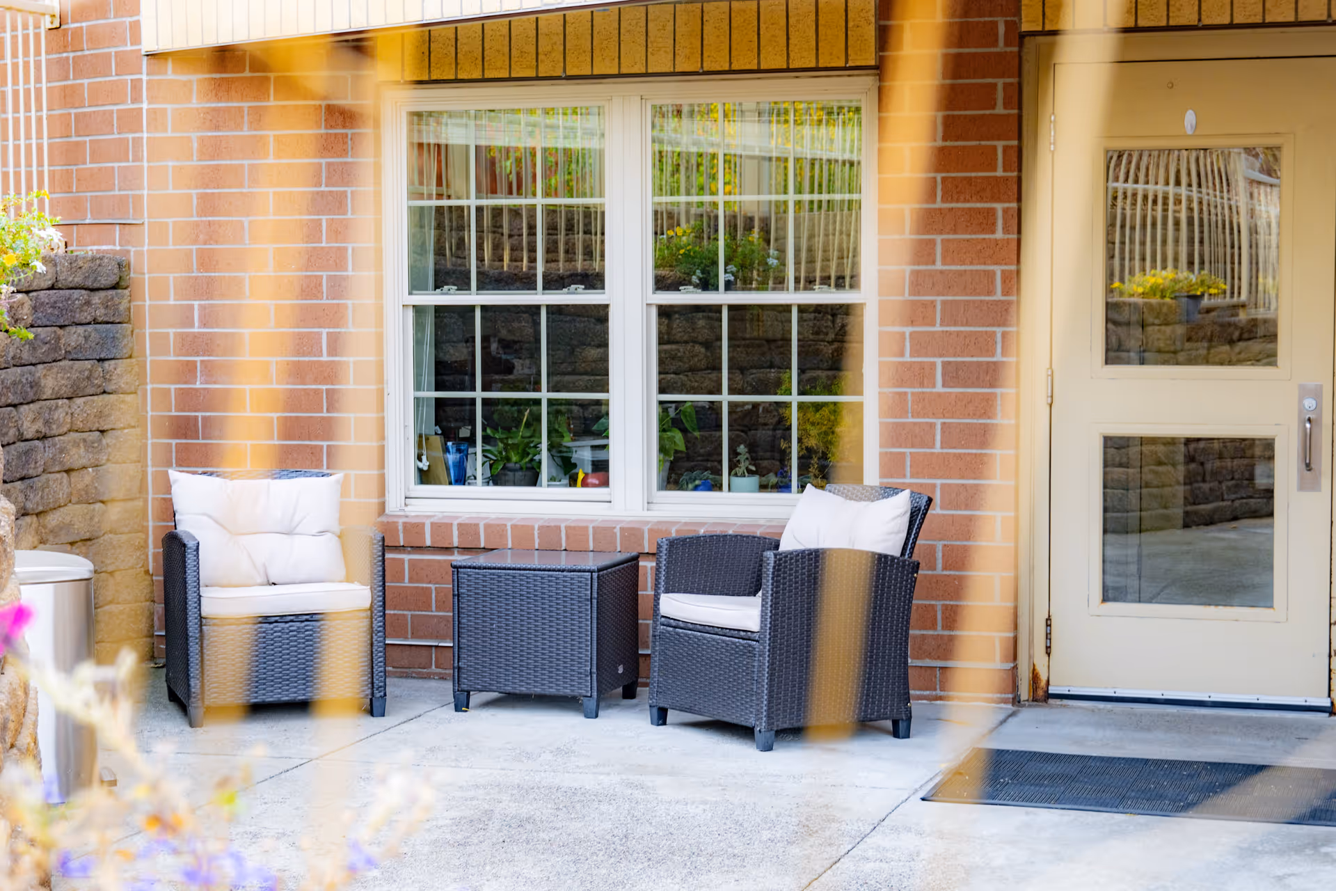 Outdoor patio area with two cushioned wicker chairs and a matching wicker table between them, set against a brick wall with a window and a door. There are plants visible through the window and a stone retaining wall to the left.