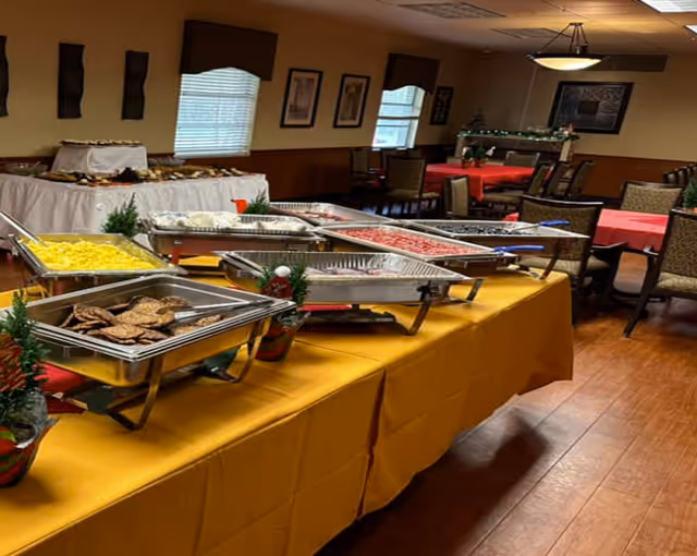 Buffet setup with trays of food including scrambled eggs and sausage patties on a yellow tablecloth in a dining room with tables and chairs arranged around the room.