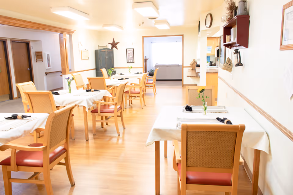 Bright dining room with several small tables and wooden chairs set with tablecloths and vases of flowers.
