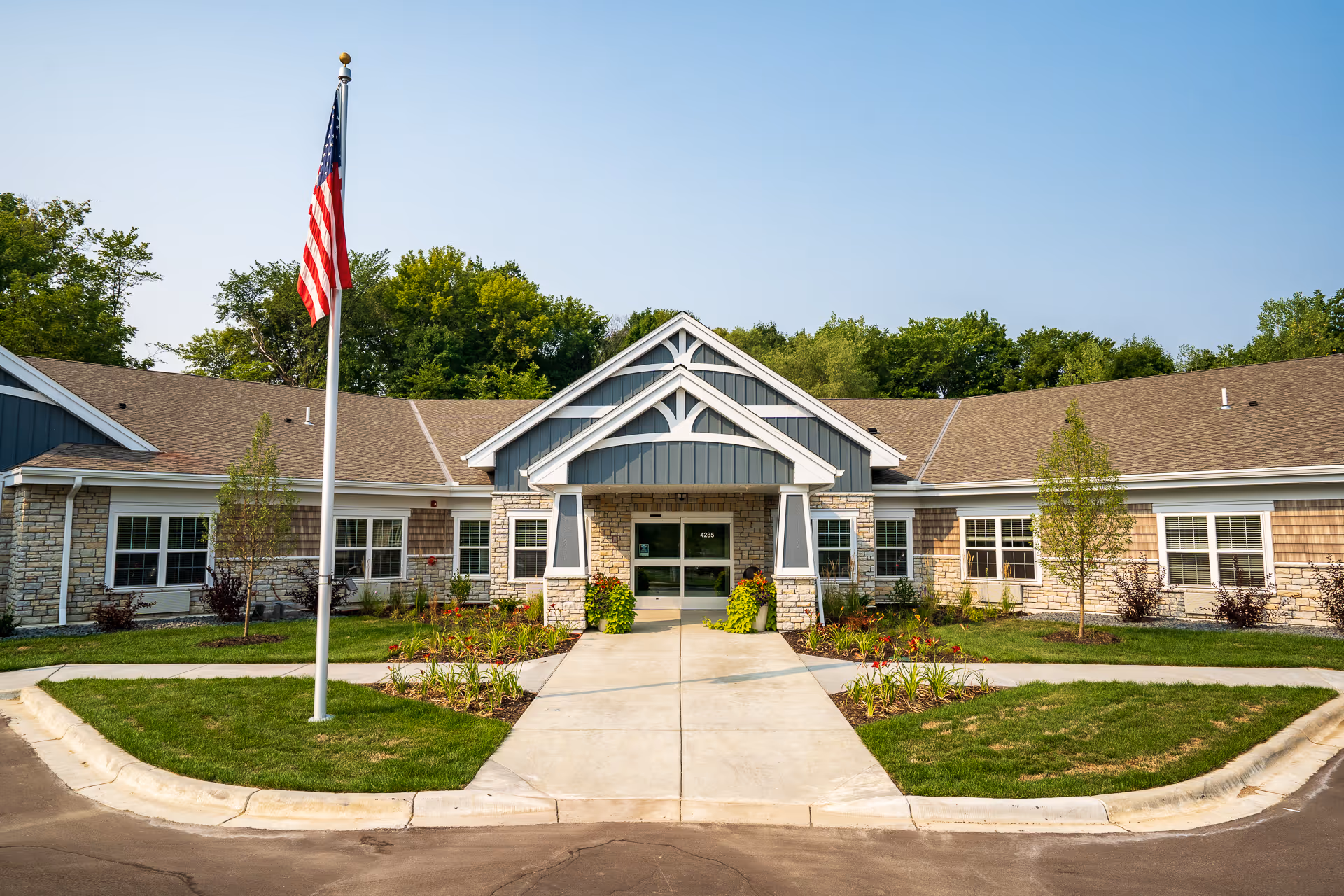 Front exterior view of NorBella Senior Living - Prior Lake building with a central entrance, stone and wood siding, a flagpole with an American flag, and landscaped greenery under a clear blue sky.