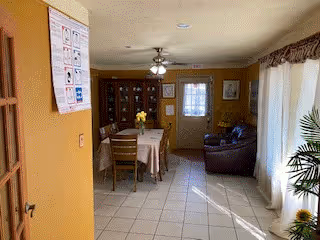 Sunlit interior showing a dining area with a table and chairs, a china cabinet and ceiling fan, with an adjacent living area featuring a sofa and windows.