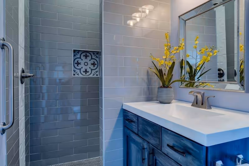 Modern bathroom with a glass-door shower, gray subway tile, a blue vanity topped with a white sink, mirror, and a vase of yellow flowers.