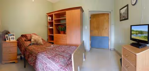 Small furnished bedroom with a single bed, dresser, bookshelf, TV, and doorway.