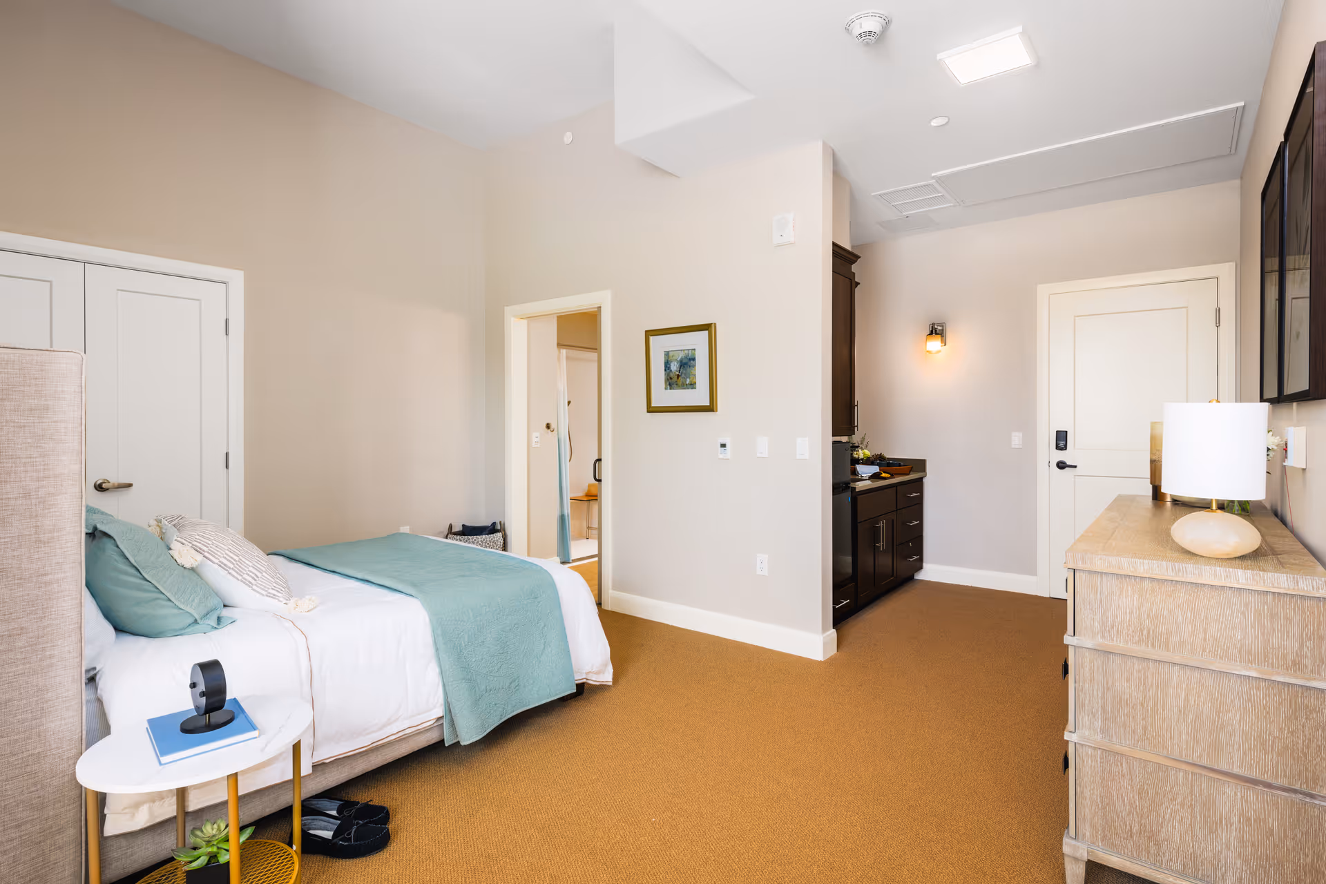 A bright and clean bedroom in a senior living facility with a bed made up with white linens and teal pillows and blanket. There is a small round side table with a book and a small camera on it, slippers on the floor, a dresser with a lamp, and a doorway leading to a bathroom. The walls are light beige and the carpet is a warm brown color.