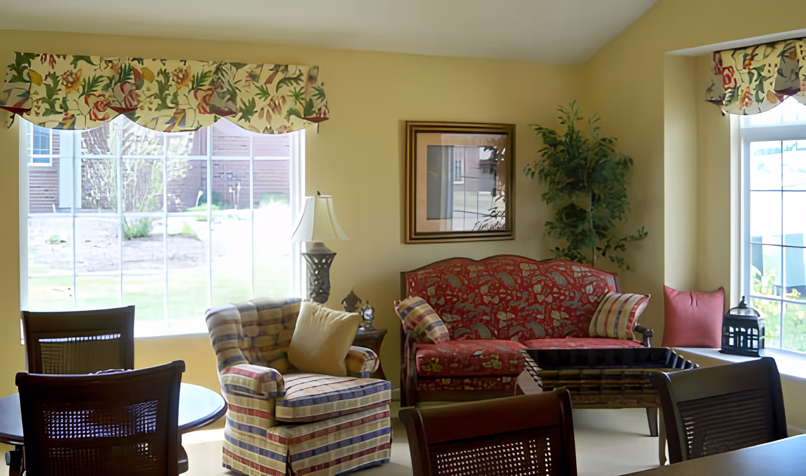 A cozy living room with a red patterned sofa, a striped armchair with a beige cushion, a wooden coffee table, and a side table with a lamp. The room has large windows with floral valances, a framed picture on the wall, and a tall green plant in the corner.