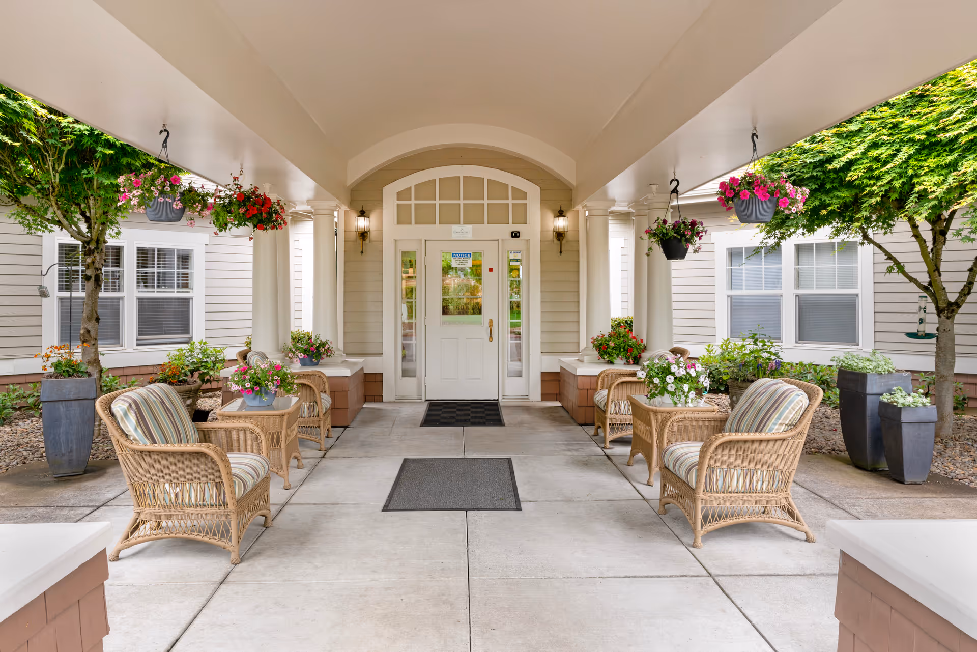 Covered outdoor seating area at Brookdale Salem with wicker chairs and small tables arranged on a concrete floor. Hanging flower pots and potted plants decorate the space, with trees and windows of the building visible on either side. A white door with glass panels is centered at the back under an arched ceiling.