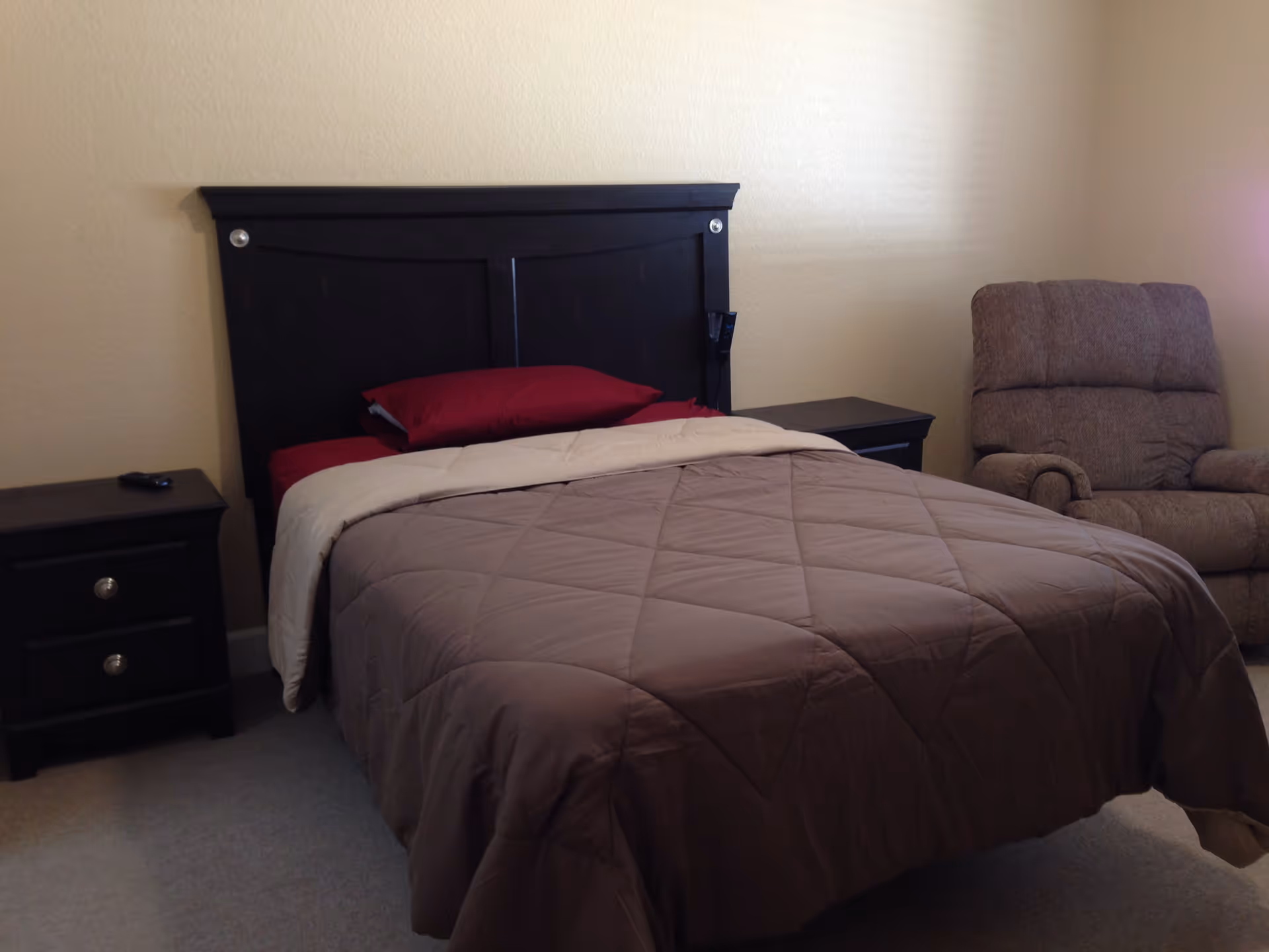 A bedroom with a dark wooden bed frame, a bed made with brown and beige bedding and red pillows, two matching dark wooden nightstands on either side of the bed, and a cushioned armchair in the corner against a beige wall.