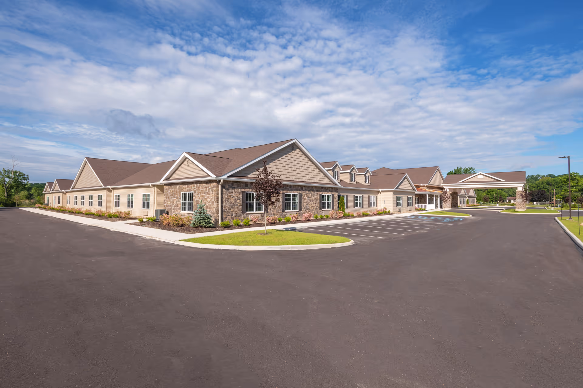 Exterior view of a single-story health and rehab facility building with beige siding and stone accents, surrounded by a large paved parking lot and landscaped greenery under a partly cloudy sky.