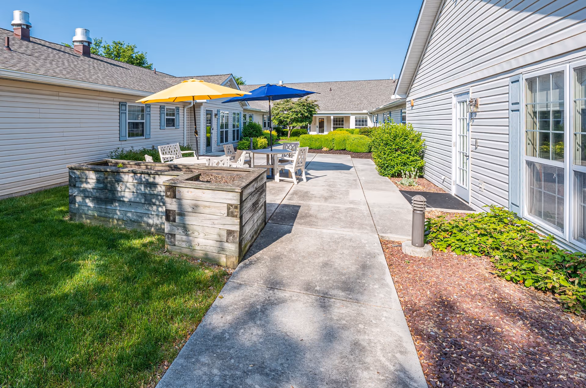 Outdoor patio area between buildings with concrete walkway, wooden raised garden beds, white chairs and tables shaded by yellow and blue umbrellas, surrounded by green bushes and grass under a clear blue sky.