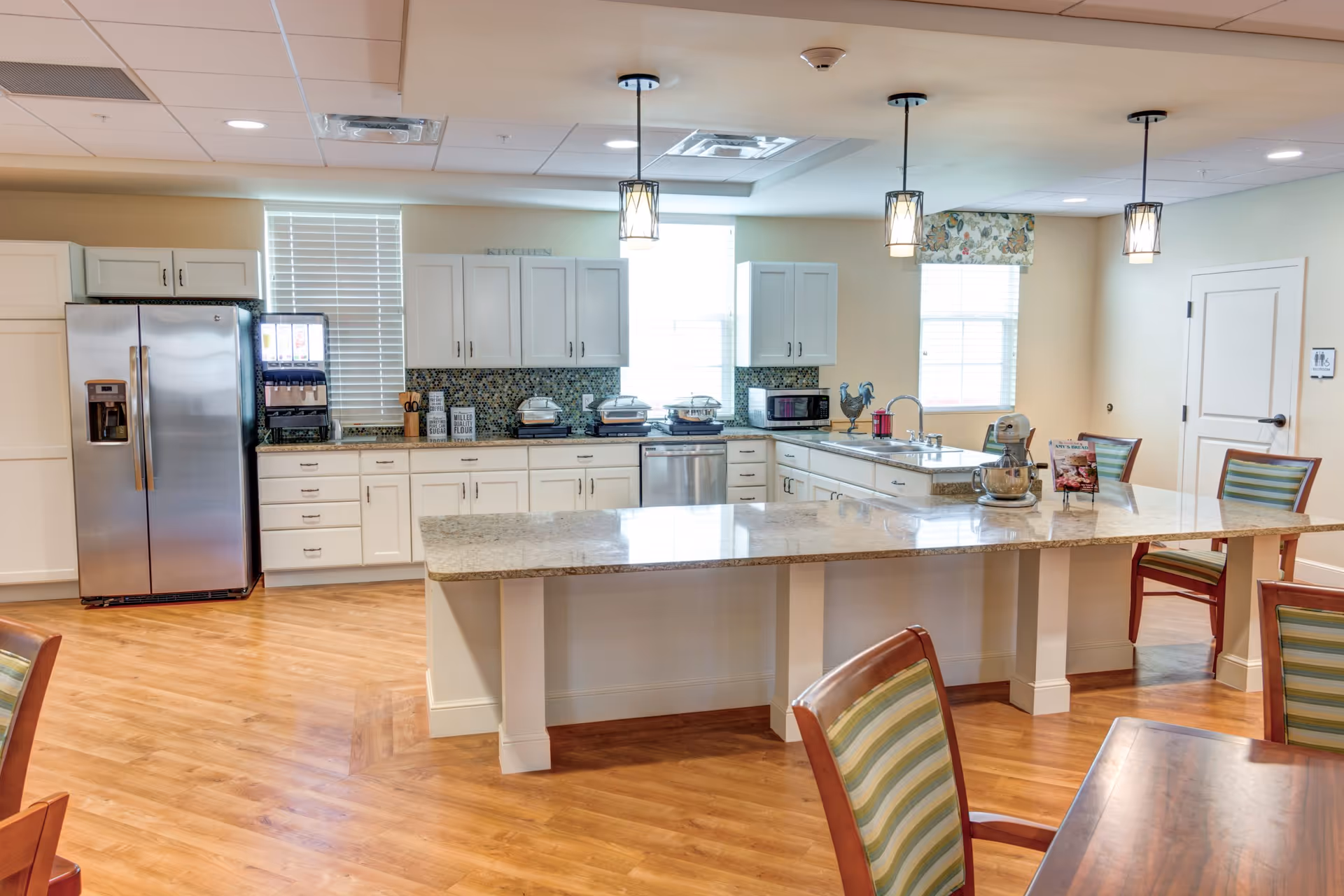 A bright and spacious kitchen area in a senior living facility with white cabinets, a large granite countertop island, stainless steel refrigerator, dishwasher, microwave, and various kitchen appliances. The room has wooden flooring, pendant lights hanging from the ceiling, and several windows with blinds allowing natural light to enter. There are also striped chairs around the island and a dining table partially visible in the foreground.
