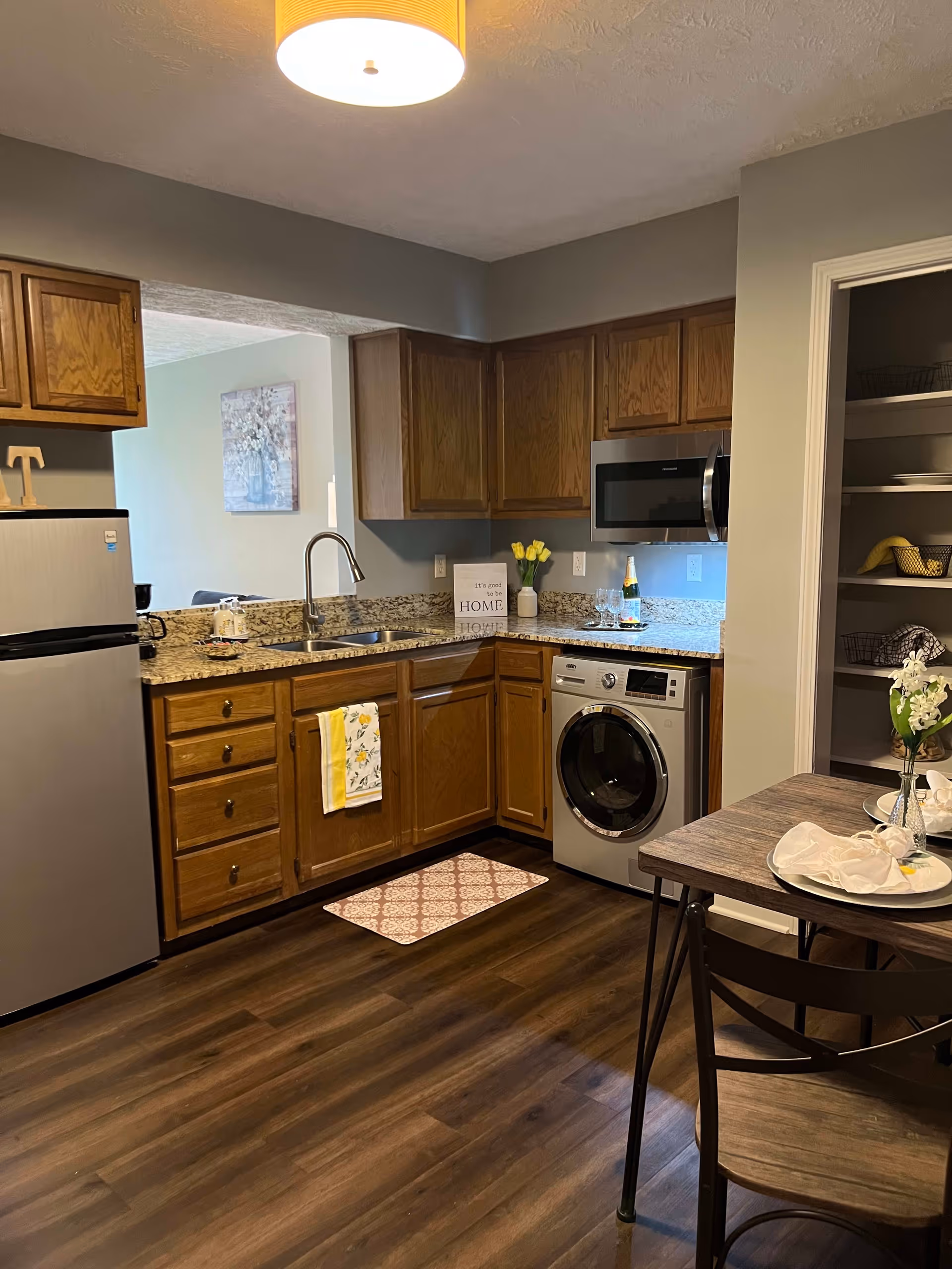 A cozy kitchen area with wooden cabinets, granite countertops, a stainless steel refrigerator, microwave, and a front-loading washing machine. There is a sink with a modern faucet, a small dining table with two chairs, and decorative items including a vase with yellow flowers and a sign that reads 'It's good to be HOME'. The floor is dark wood, and there is a light fixture hanging from the ceiling.