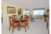 A bright and spacious living and dining area in a senior living community. The dining area features a round wooden table with four chairs that have red cushioned seats. A chandelier hangs above the table. The living area includes a rocking chair, a sofa, a side table with a lamp, and a large window with curtains allowing natural light to fill the room. The walls are decorated with framed artwork and the floor is carpeted in a light color.