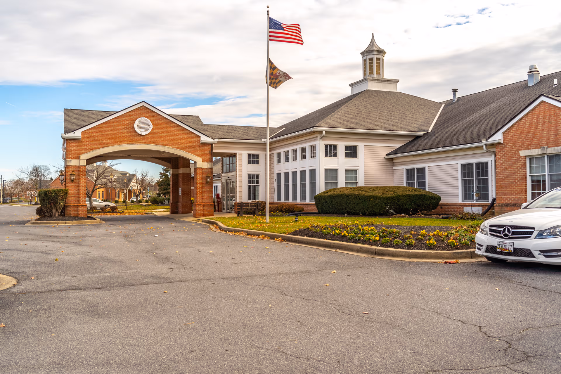 Exterior view of Autumn Lake Healthcare at Glade Valley showing a brick and siding building with a covered entrance, an American flag and a Maryland state flag on a flagpole, landscaped flower beds, and a white Mercedes-Benz car parked on the right.