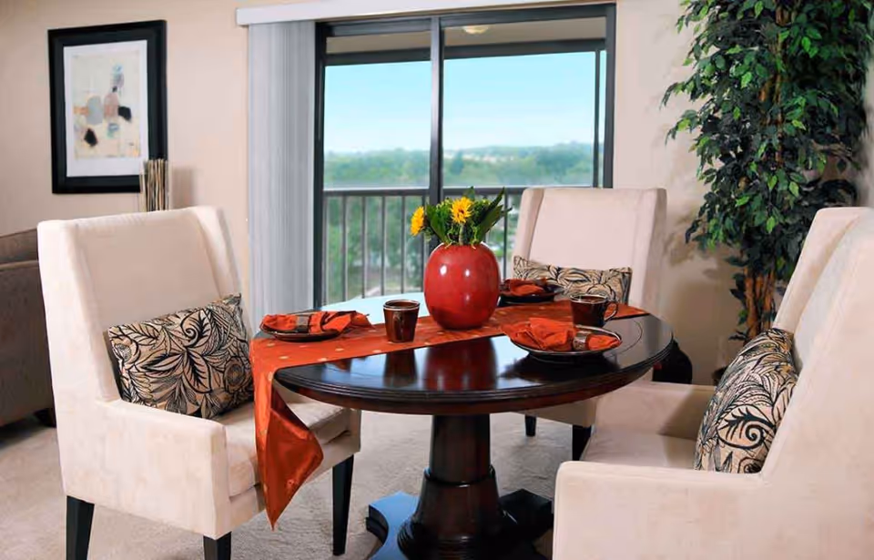 Round wooden dining table set for four with beige upholstered chairs, a red runner and vase, in a bright interior with sliding glass doors overlooking a balcony.