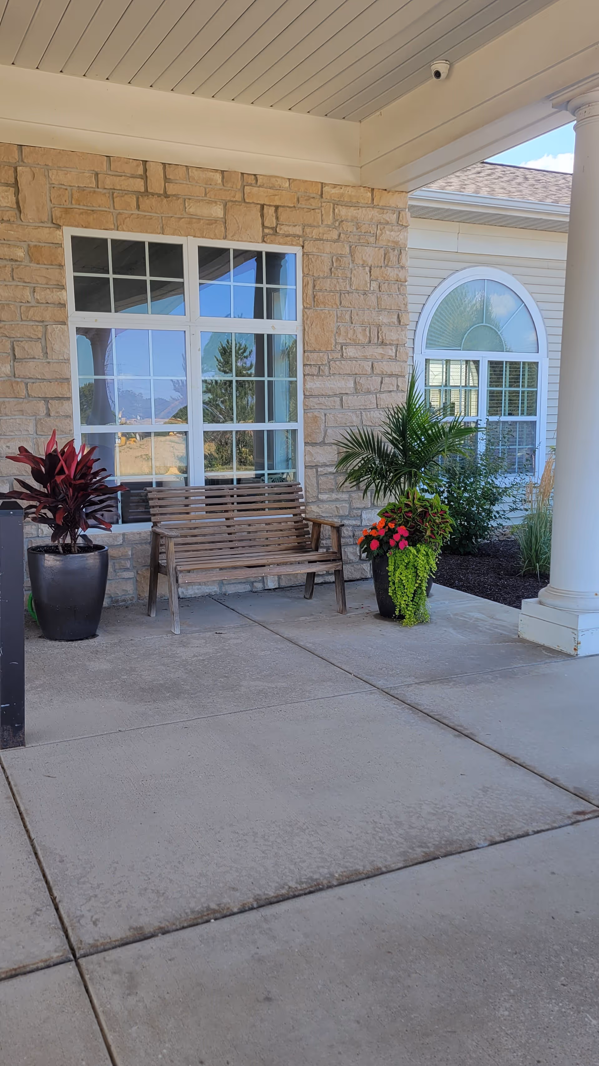 Covered outdoor seating area with a wooden bench placed against a stone wall. Two large potted plants with green and red foliage flank the bench. A large window with white framing is behind the bench, and a white column supports the roof of the covered area. Another window with an arched top is visible on the adjacent wall.
