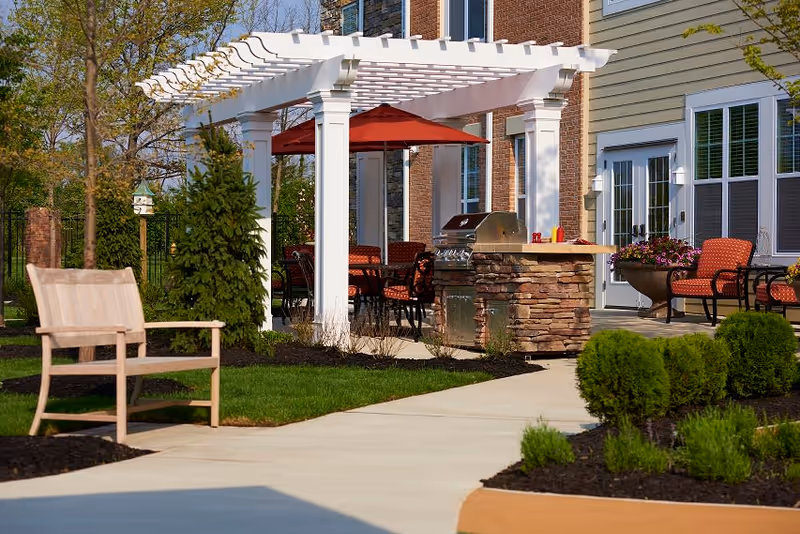 Outdoor patio area at Dublin Retirement Village featuring a white pergola with a red umbrella underneath, outdoor seating with red cushions, a stone grill station, a wooden bench, and landscaped greenery along a paved walkway.