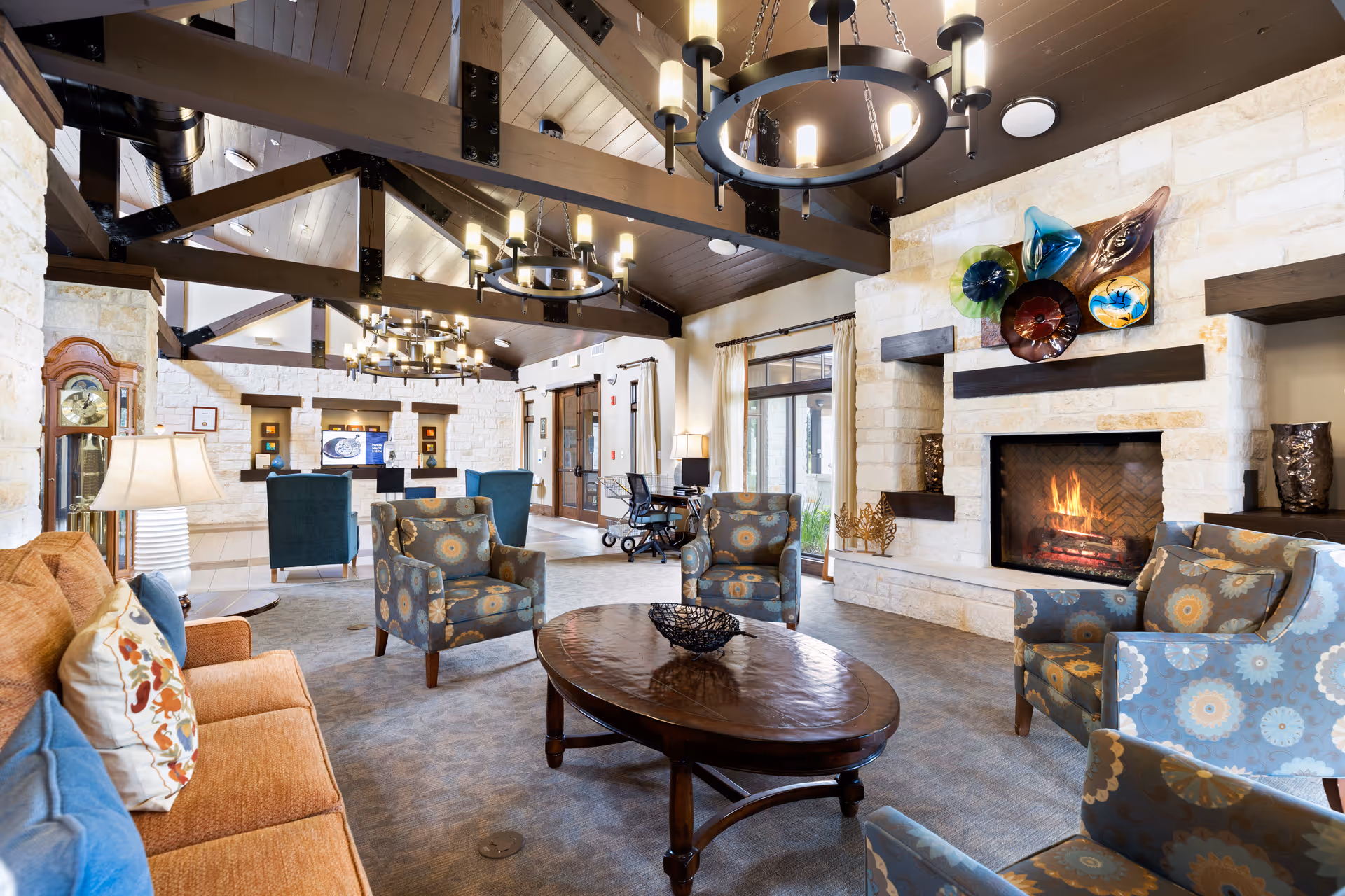 A spacious and well-lit living room area with a high wooden beam ceiling and multiple circular chandeliers. The room features a stone fireplace with a fire burning, decorated with colorful glass art above it. There are several patterned armchairs and a brown sofa with decorative pillows arranged around a wooden coffee table. The room has large windows with curtains, a grandfather clock, and a reception desk area in the background.