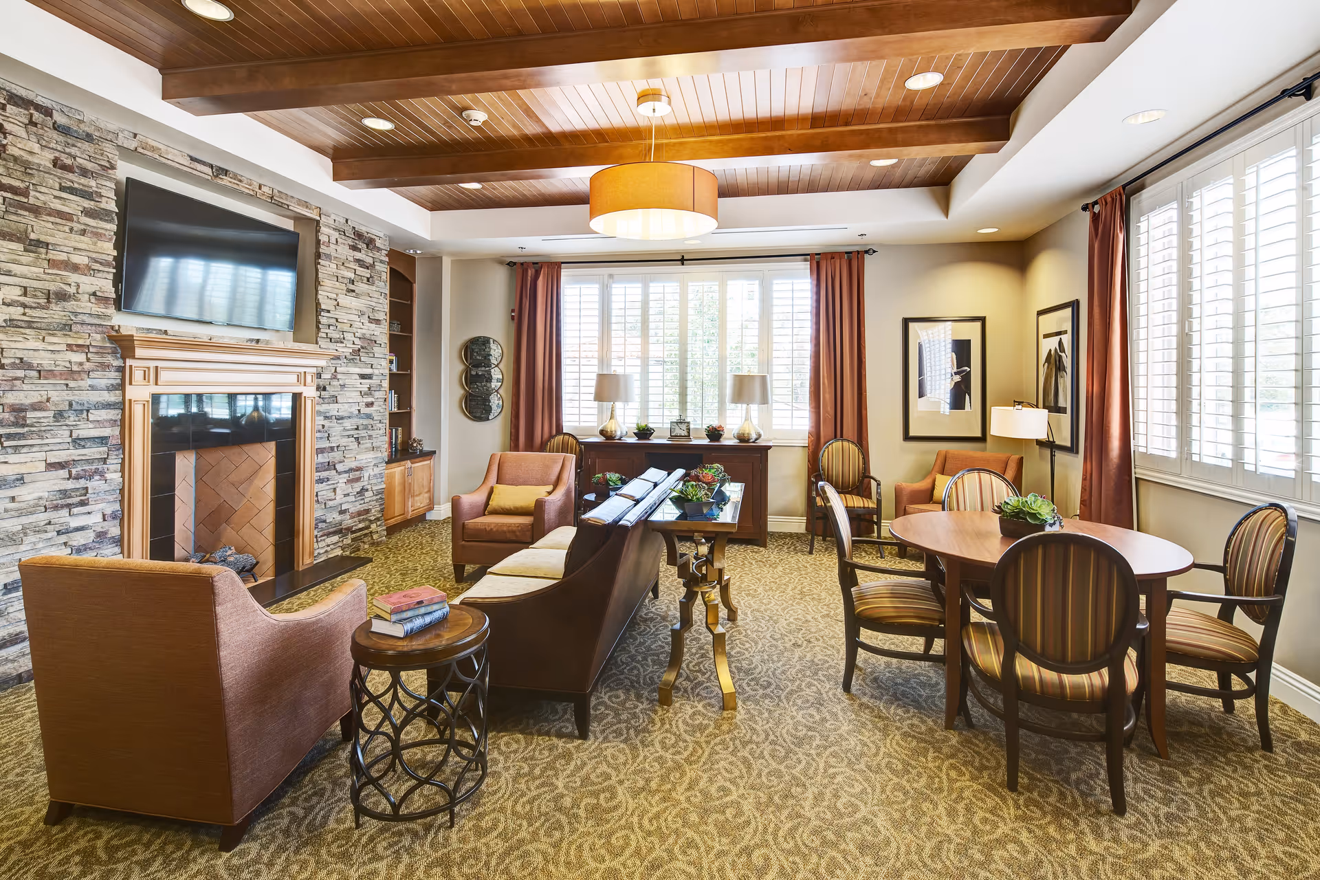 Well-appointed communal living room with upholstered chairs, a sofa, fireplace and TV, and a round table by large windows.