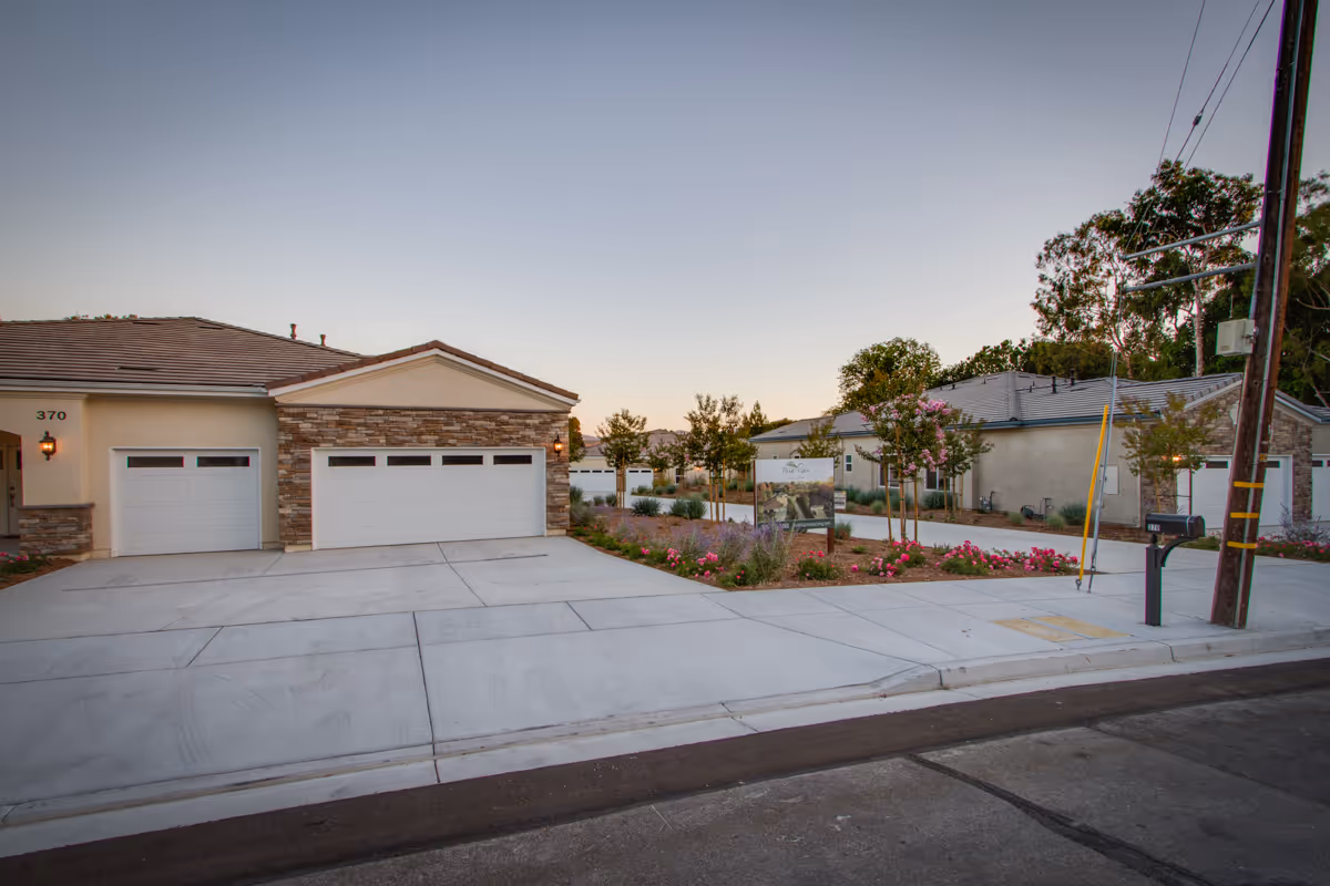 Exterior view of a single-story senior living building with three garage doors, paved driveways, and landscaped flower beds.