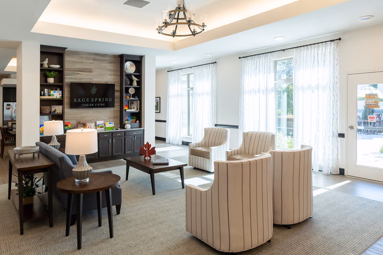 Bright and spacious senior living common area with large windows covered by sheer white curtains, four striped armchairs arranged around a wooden coffee table, a gray sofa with side tables and lamps, and a built-in shelving unit with a TV displaying the Sage Spring Senior Living logo.