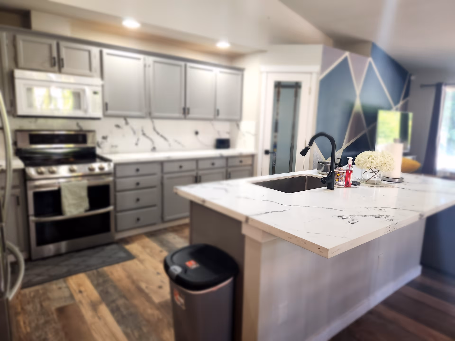 Modern kitchen with gray cabinets, stainless steel stove and microwave, white marble countertops, a kitchen island with a black faucet and sink, a vase with white flowers, hand soap, and a trash can on wooden flooring.