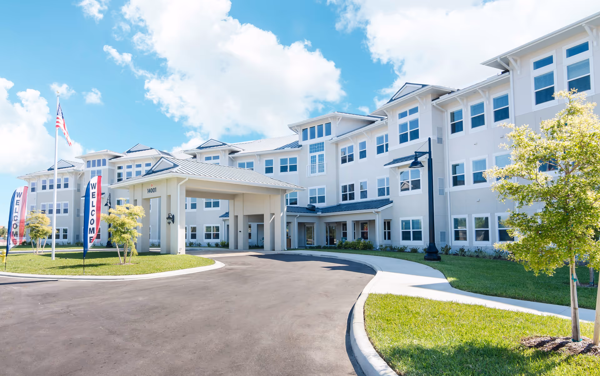 Exterior view of a large, modern three-story senior living facility building with a covered entrance, a curved driveway, green lawns, small trees, an American flag, and two vertical banners that say 'WELCOME' under a partly cloudy blue sky.