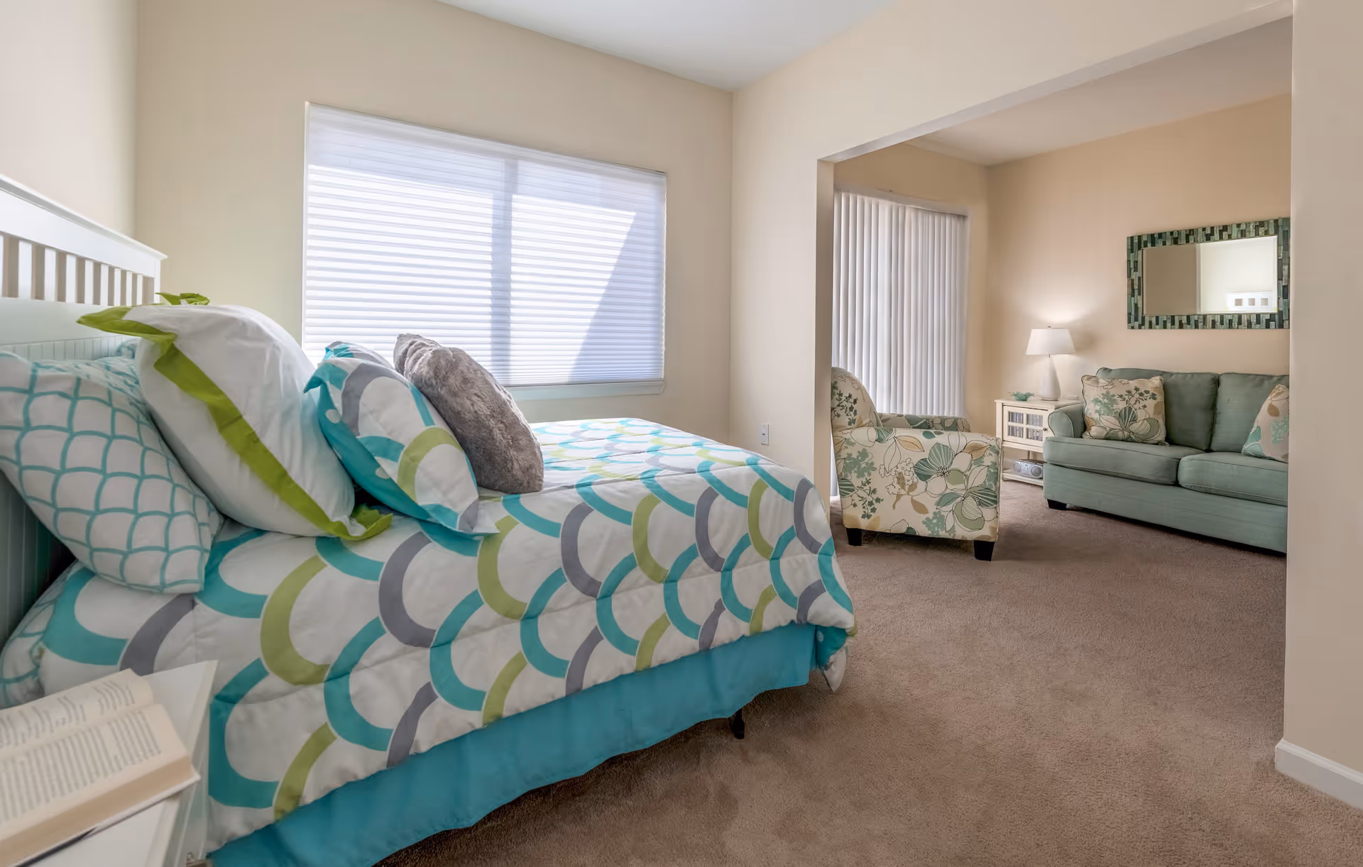 A bright bedroom with a bed covered in a colorful patterned comforter and multiple pillows. Next to the bed is a small white nightstand with an open book. The room has a large window with blinds letting in natural light. Through an open doorway, a cozy living area is visible with a floral armchair, a green sofa with patterned cushions, a white side table with a lamp, and a decorative mirror on the wall.