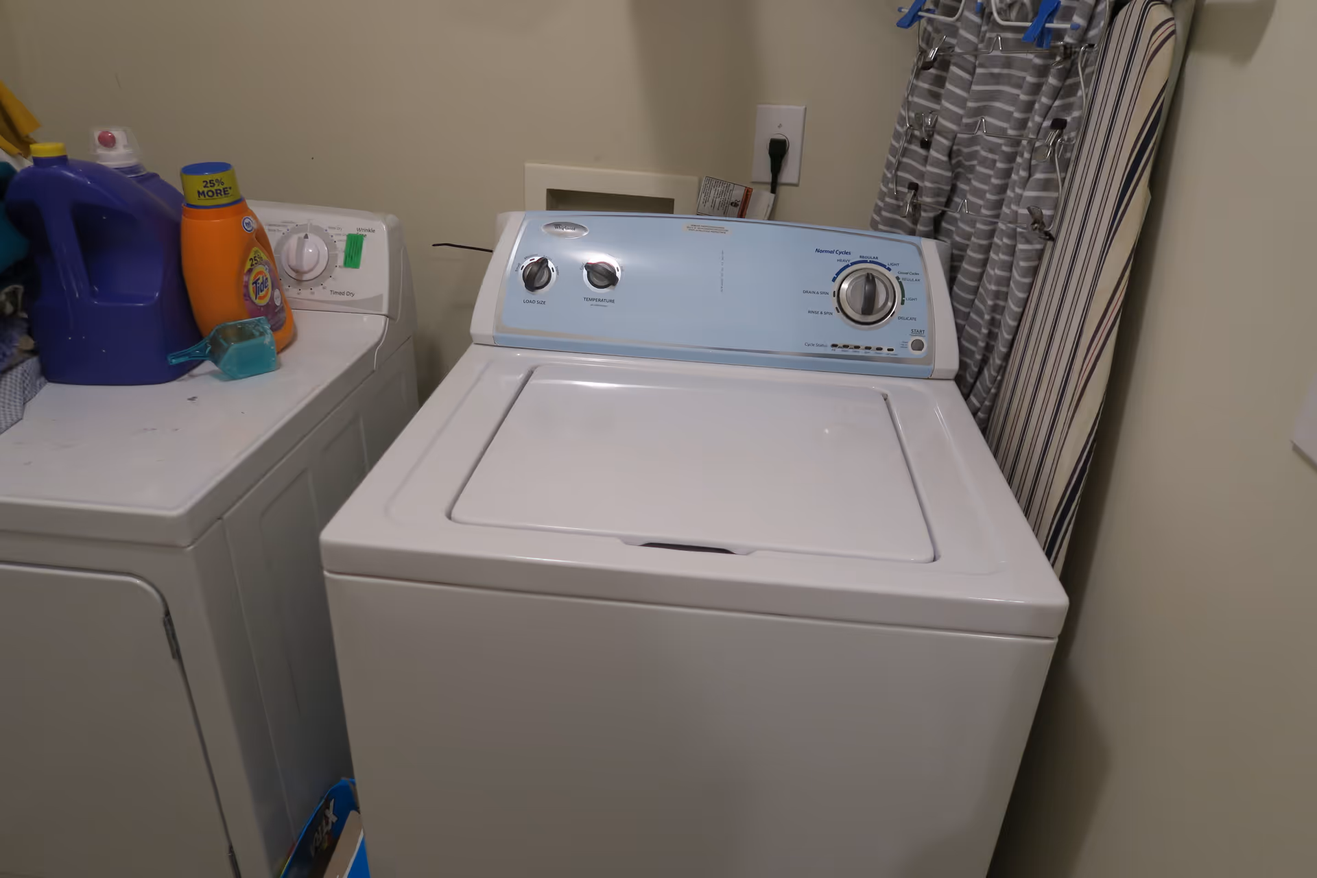 A laundry room with a white washing machine and a white dryer side by side. On top of the dryer are several laundry detergent bottles, including a large purple bottle and an orange Tide bottle. An ironing board with a striped cover and a gray hanging organizer with clips are visible leaning against the wall.