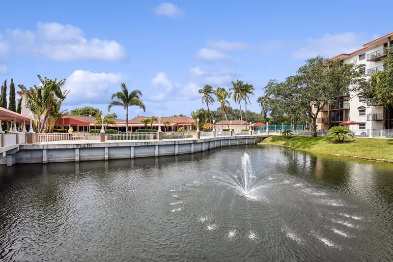 Outdoor view of a senior living facility named Fountainview featuring a large pond with a central water fountain, surrounded by green grass, palm trees, and buildings with balconies under a partly cloudy blue sky.