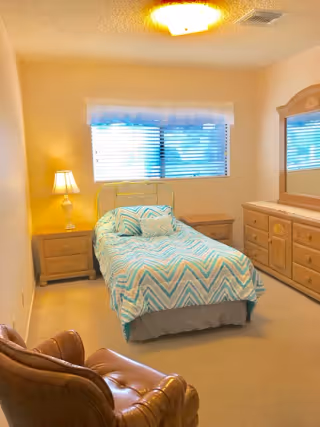 A small bedroom with a single bed covered in a blue and white chevron patterned bedspread and matching pillows. There is a wooden nightstand with a lamp on the left side of the bed, a wooden dresser with a mirror on the right wall, and a brown leather armchair in the foreground. A window with blinds and a sheer curtain is above the bed, letting in natural light.