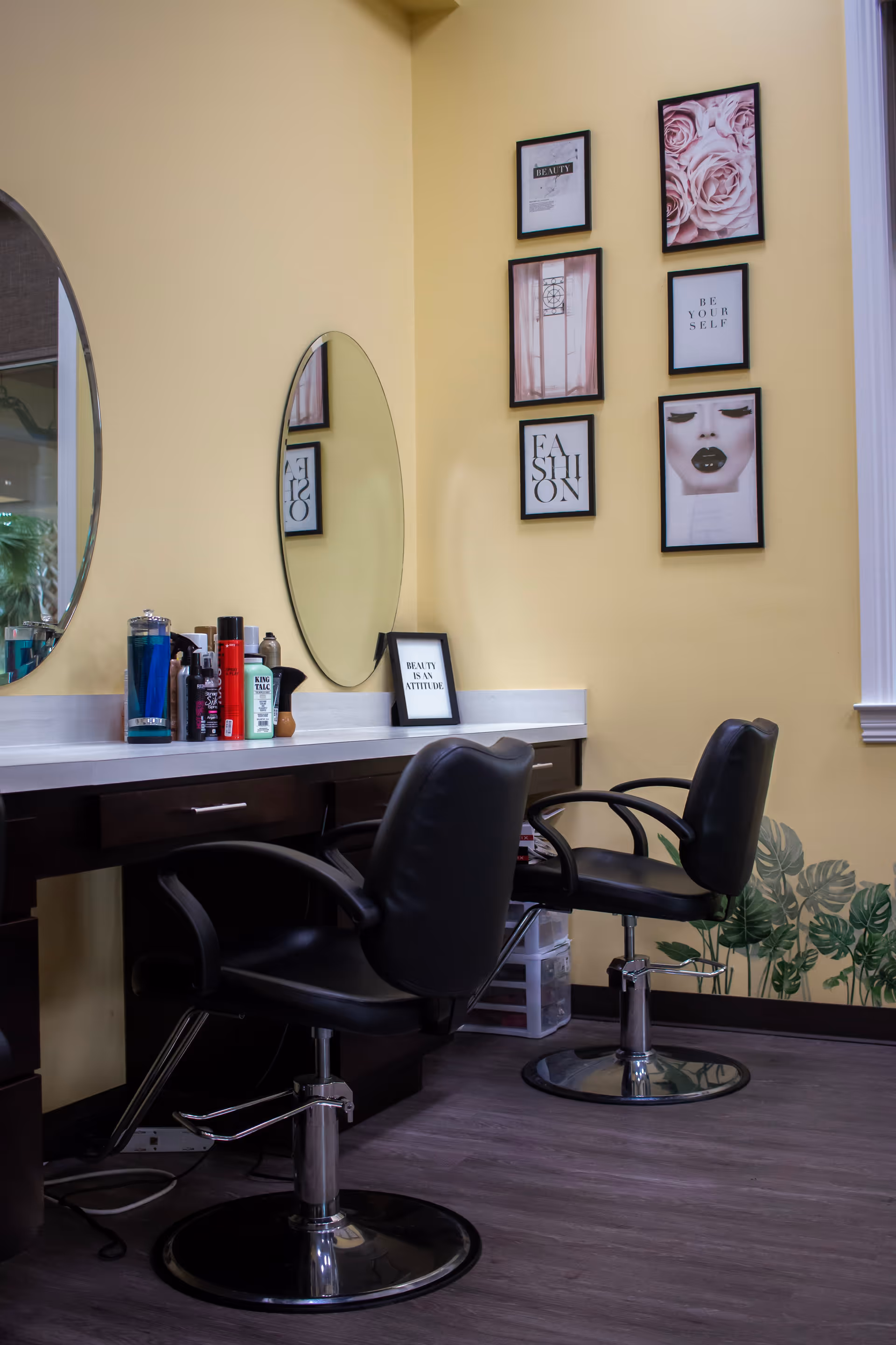 Interior view of a salon area with two black salon chairs in front of a counter with various hair and beauty products. Two oval mirrors are mounted on a yellow wall, which is decorated with framed pictures and motivational quotes. There is a window on the right side and a plant decal near the floor.