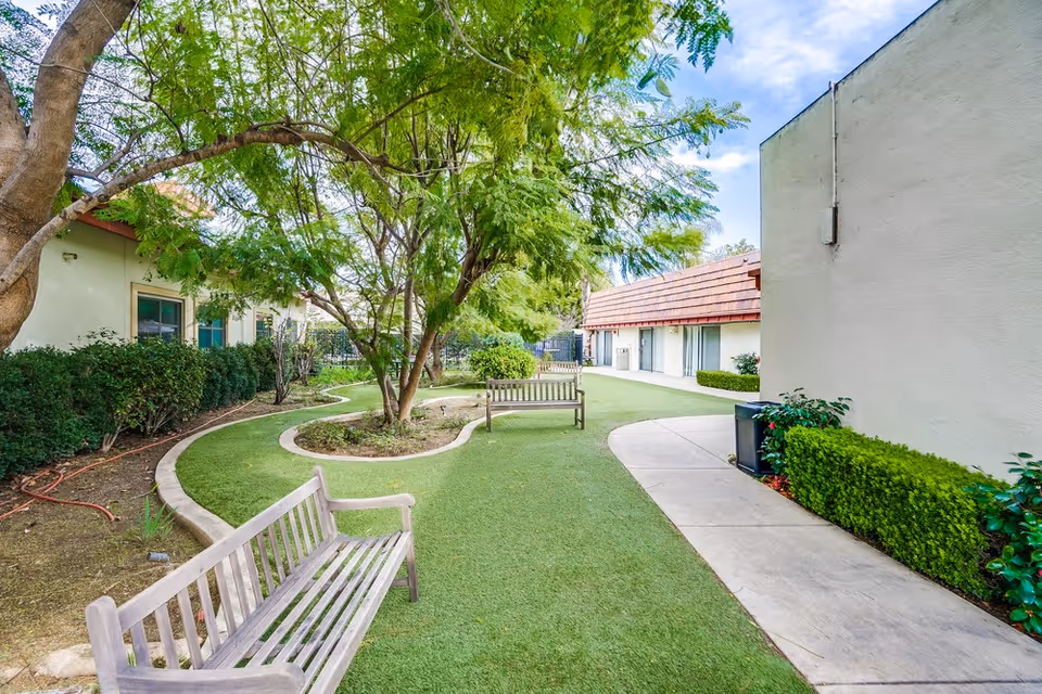 A landscaped courtyard at a care facility with benches, trees, manicured lawn, and paved walkways between low buildings.