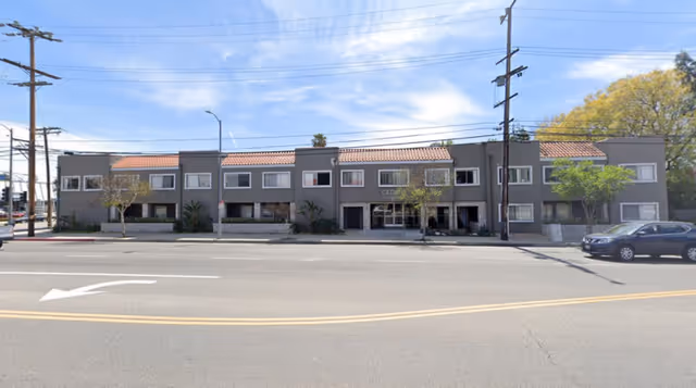 Exterior front view of a two-story assisted living facility building with a tiled roof, multiple windows, and a main entrance in the center. There are a few small trees and shrubs in front of the building, a parked car on the right side, and power lines overhead.