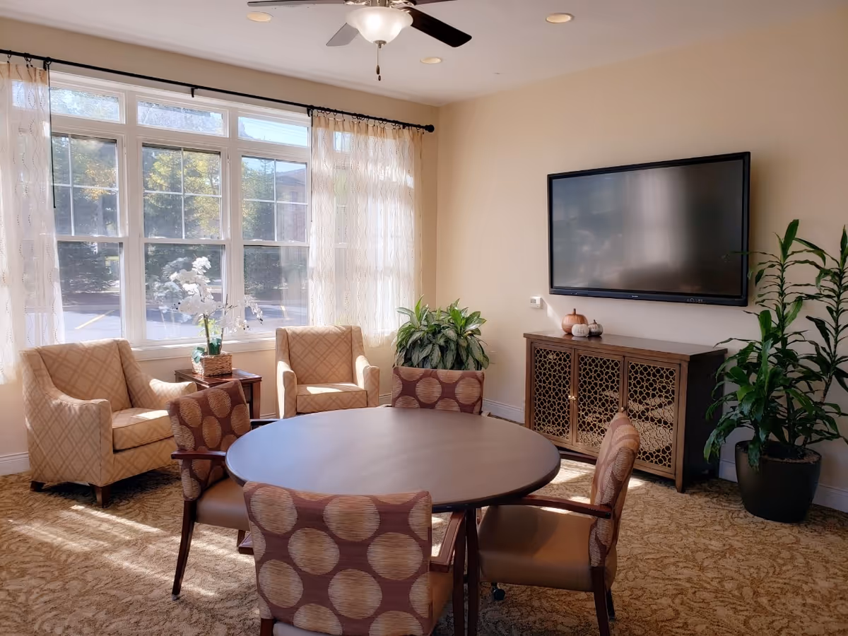 A bright room with a round wooden table surrounded by four chairs with patterned upholstery. Two beige armchairs are placed near a large window with sheer curtains, allowing natural light to fill the room. A flat-screen TV is mounted on the wall above a decorative wooden cabinet. There are two potted plants in the corners, and the room has a beige carpet and light-colored walls.