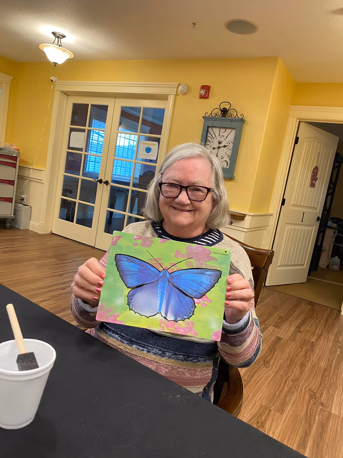 An elderly woman with gray hair and glasses is sitting at a table in a room with yellow walls and wooden flooring. She is smiling and holding up a colorful painting of a large blue butterfly on a green and pink background. On the table in front of her is a white cup with a paintbrush inside. Behind her are double glass doors and a wall clock.