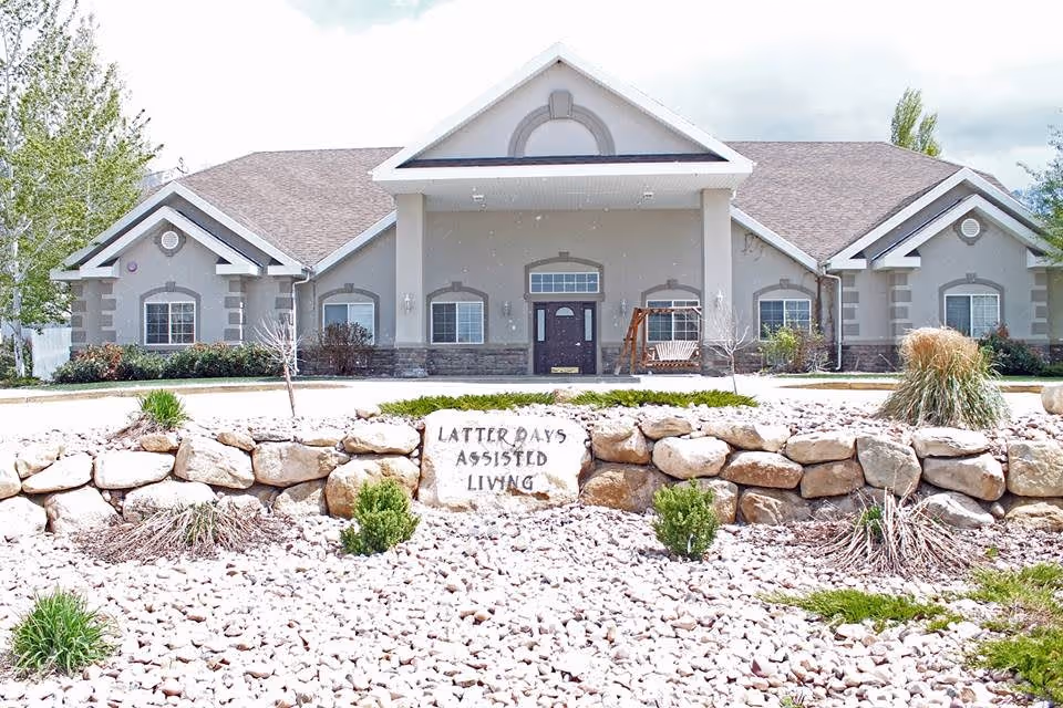 Front exterior view of a single-story assisted living facility building with a peaked roof and multiple windows. In front of the building is a landscaped area with rocks, small bushes, and a stone sign that reads 'LATTER DAYS ASSISTED LIVING'.