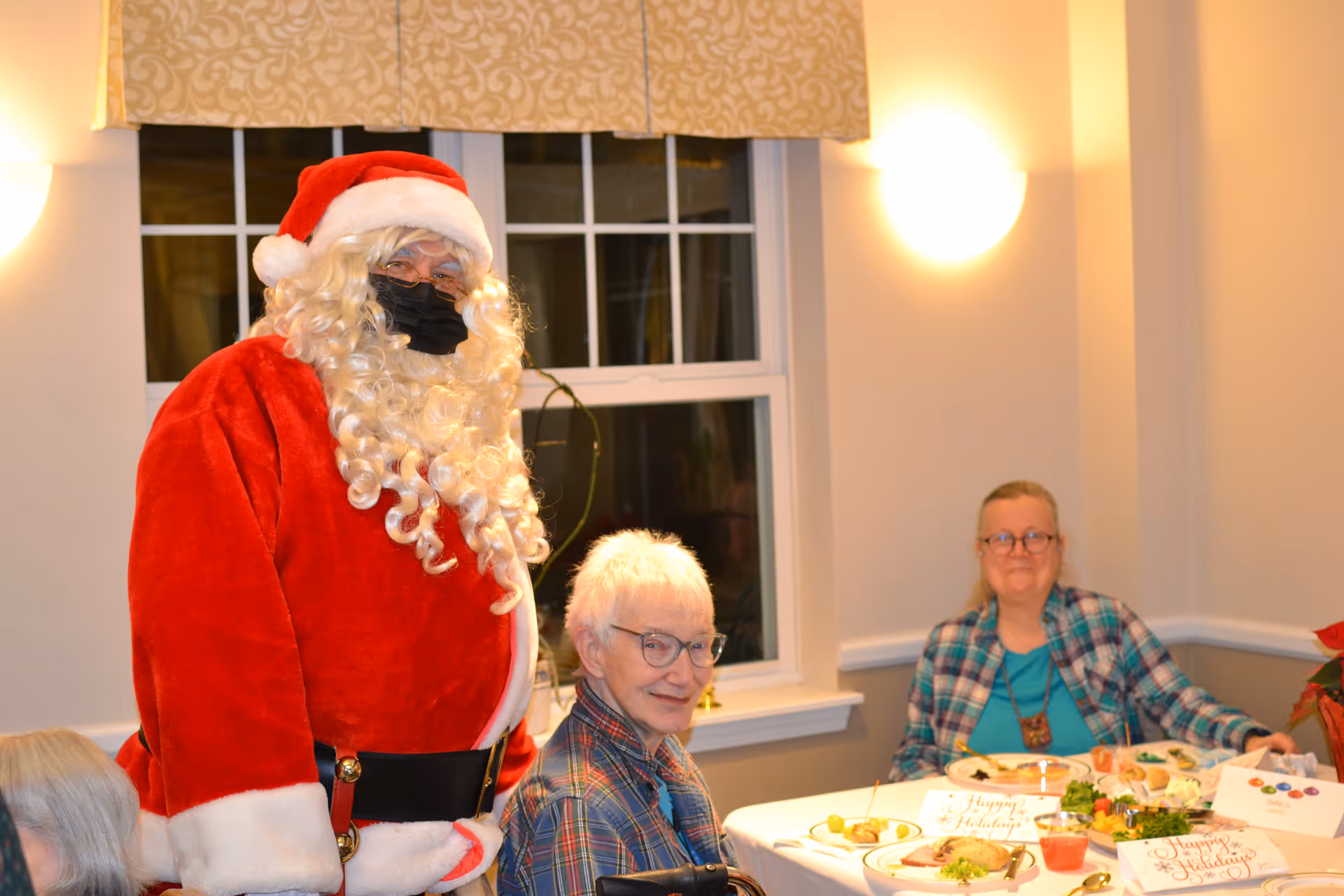 A masked Santa Claus stands beside two seated senior residents at a holiday-decorated dining table inside a dining room.