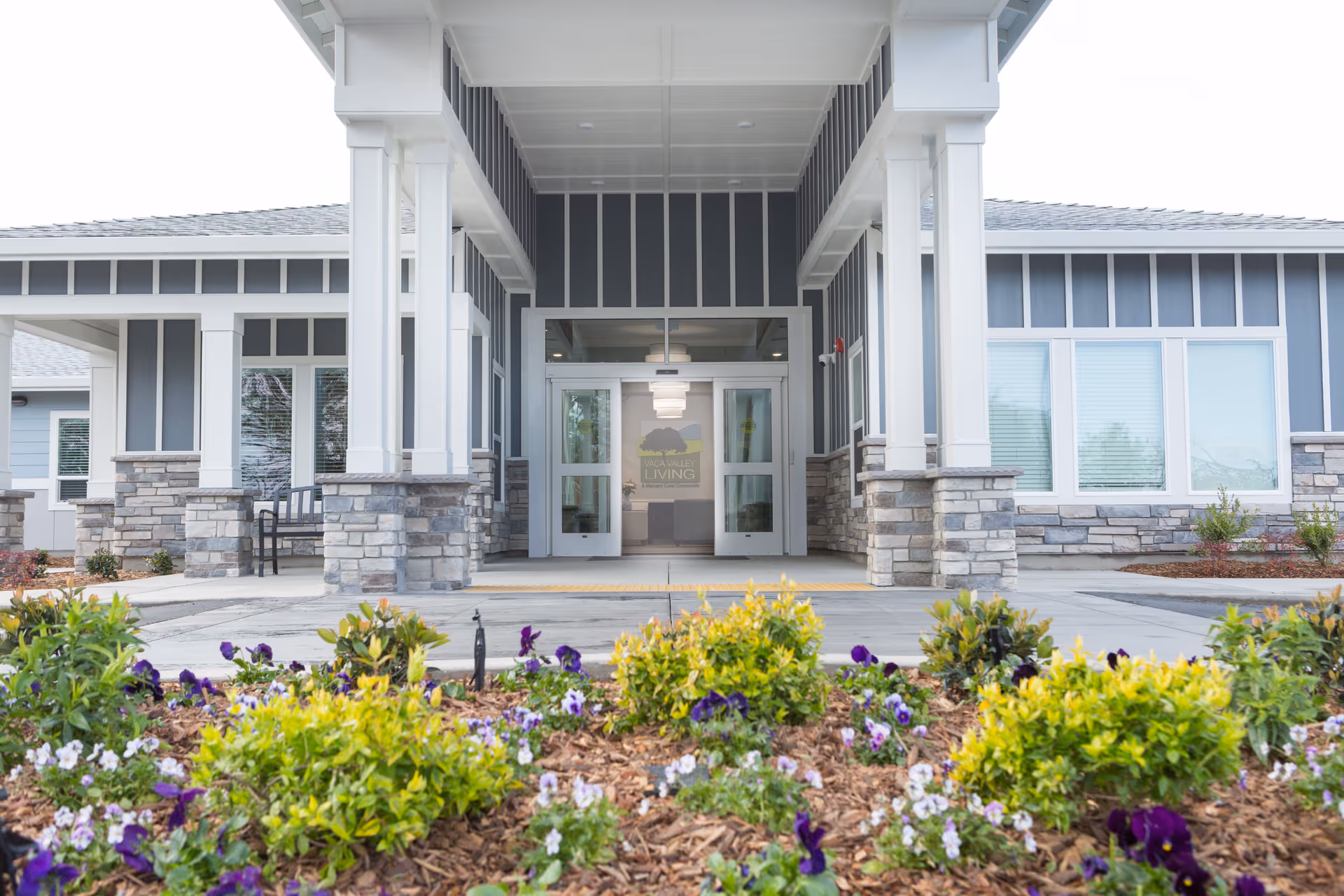 Entrance of Vaca Valley Living facility showing a covered porch with white pillars and stone bases, a pathway leading to double glass doors, and a flower bed with green shrubs and purple and white flowers in the foreground.