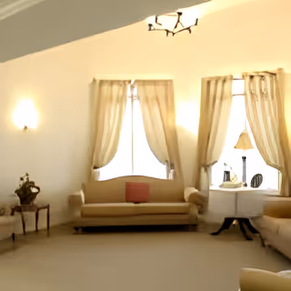 A bright living room with large windows covered by sheer curtains, two beige sofas facing each other, a round white table with a lamp and decorative items, and a small side table with a plant. The walls are painted white and the room has a beige carpet.