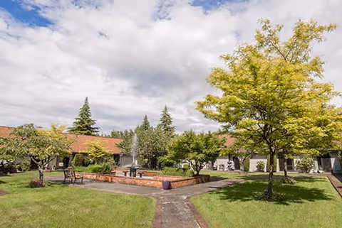 Outdoor courtyard area with green grass, several trees, a brick fountain in the center, benches, and a pathway leading through the space. The courtyard is surrounded by a single-story building with a red roof under a partly cloudy sky.
