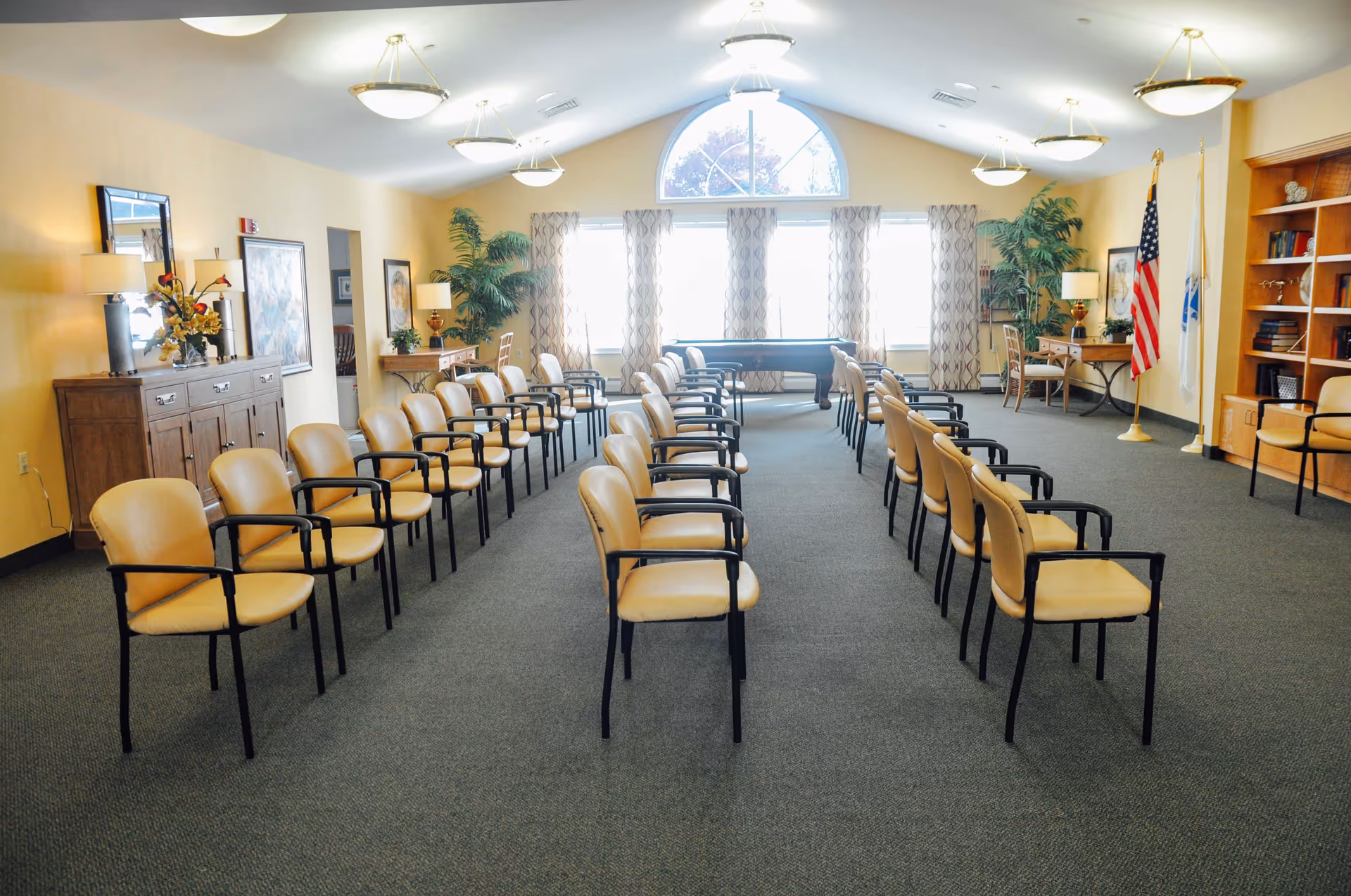A large, well-lit room with rows of beige chairs arranged in two columns facing forward. The room has a high ceiling with hanging light fixtures, a large arched window with patterned curtains, and various pieces of furniture including a wooden cabinet, desks with lamps, potted plants, and bookshelves. There are also two flags positioned near the right side of the room.