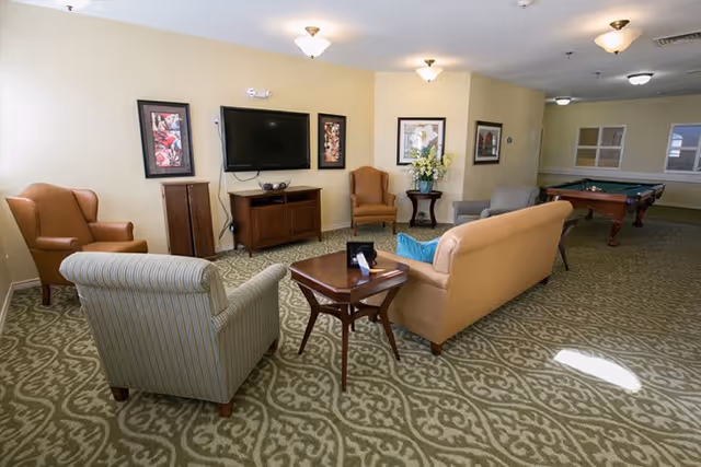 A senior living facility common area with a patterned carpet, several upholstered chairs and a sofa arranged around a wooden coffee table, a flat-screen TV mounted on the wall, framed artwork, and a pool table in the background.