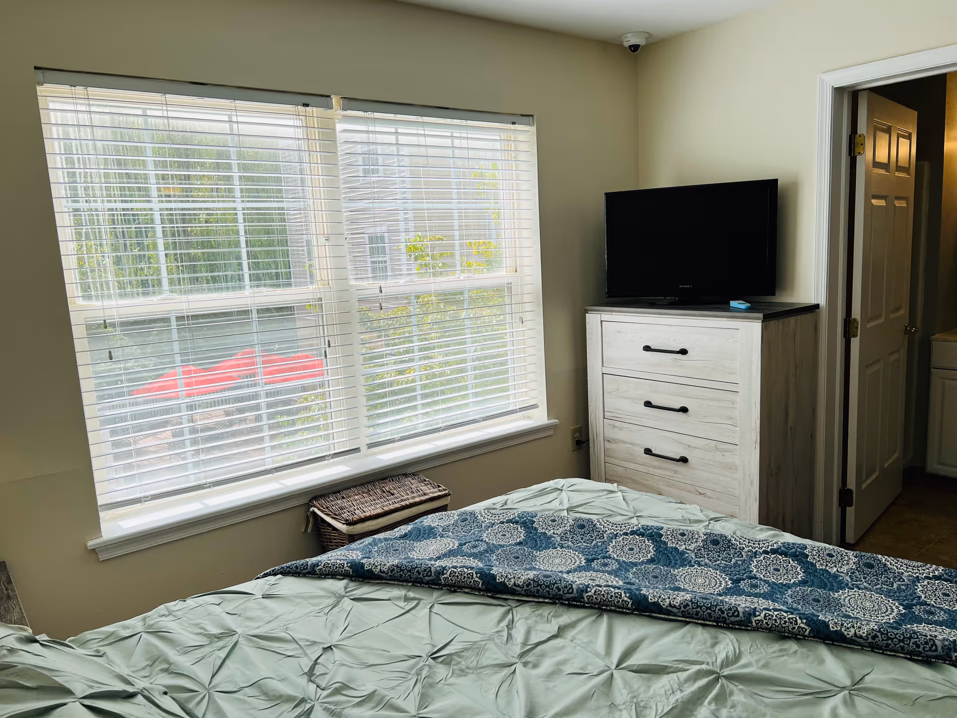 Bright bedroom with a bed in the foreground, a large window with blinds, a dresser topped by a TV, and a doorway to an en-suite bathroom.