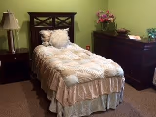 A single bed with a wooden headboard and a beige patterned comforter in a room with light green walls. Next to the bed is a nightstand with a lamp, and across from the bed is a dark wooden dresser with a vase of flowers and other decorative items on top.