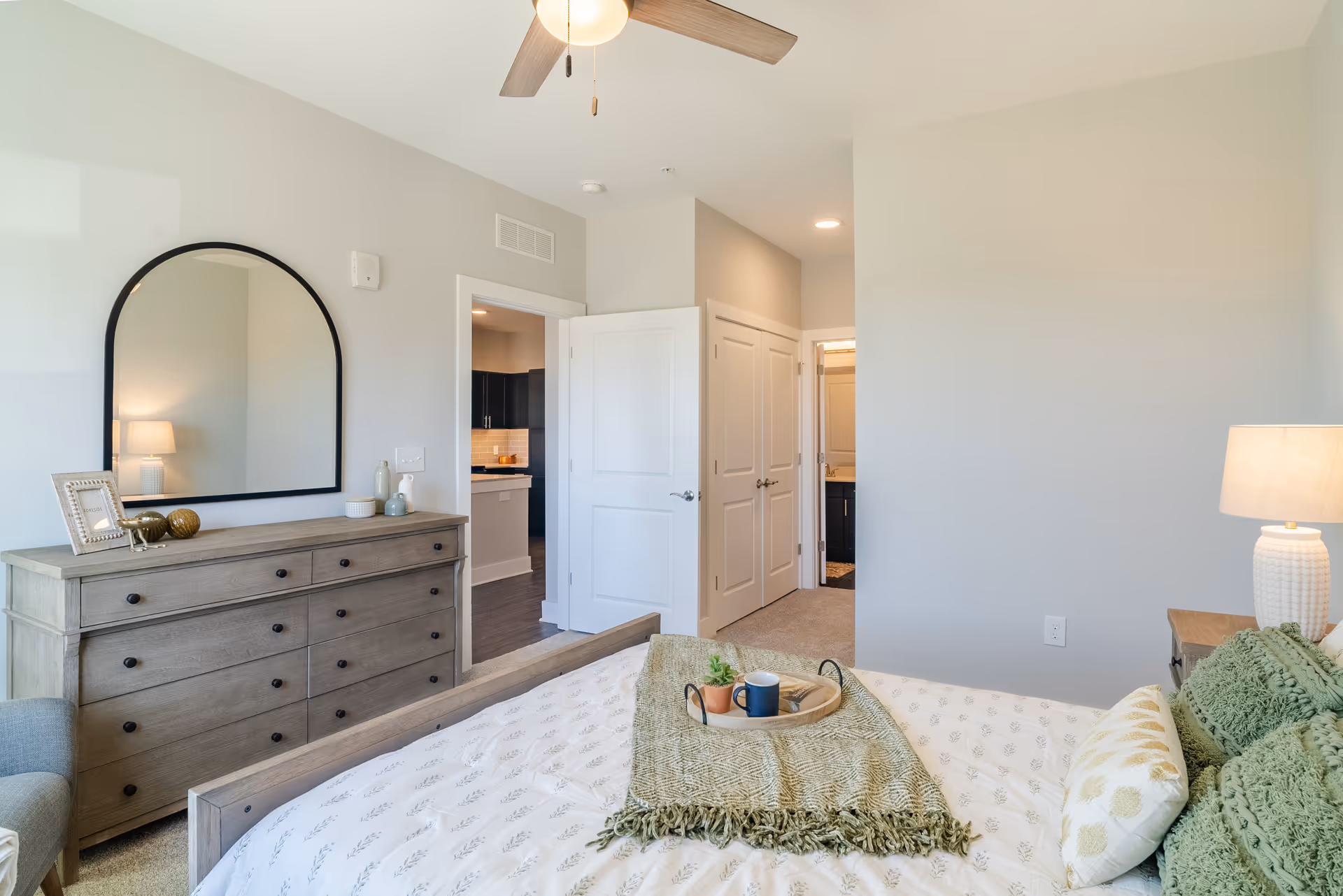 A bright bedroom with a bed covered in white and green bedding, a wooden dresser with a large arched mirror above it, and a lamp on a nightstand. The room has light-colored walls and carpeted floor. There are open doors leading to a kitchen area and a bathroom.