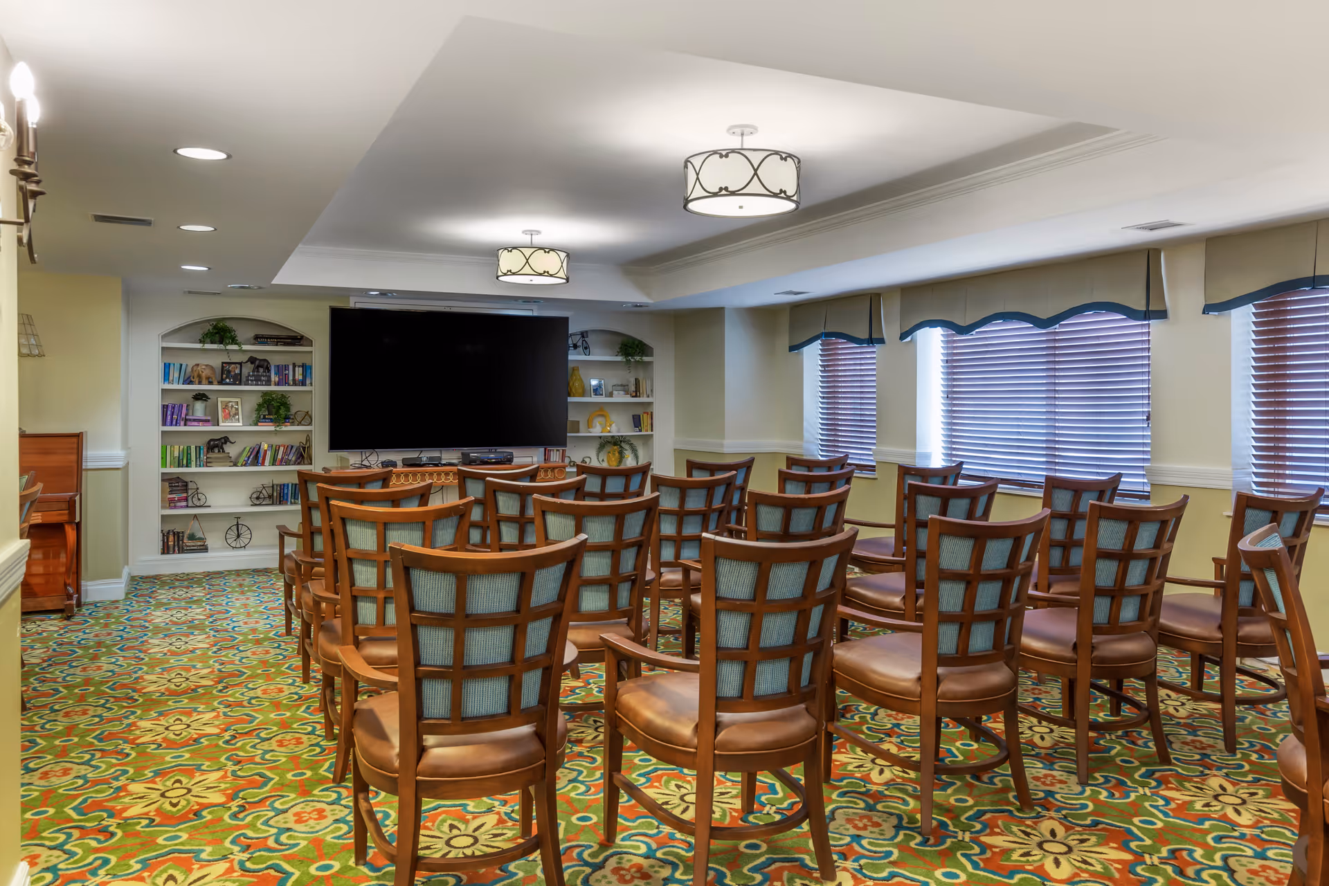 A community room with rows of wooden chairs facing a large wall-mounted TV and built-in bookshelves.