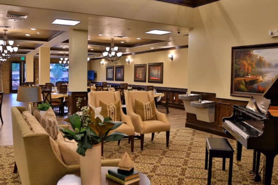 Spacious senior living common area with lounge seating, dining tables, chandeliers, and a grand piano.