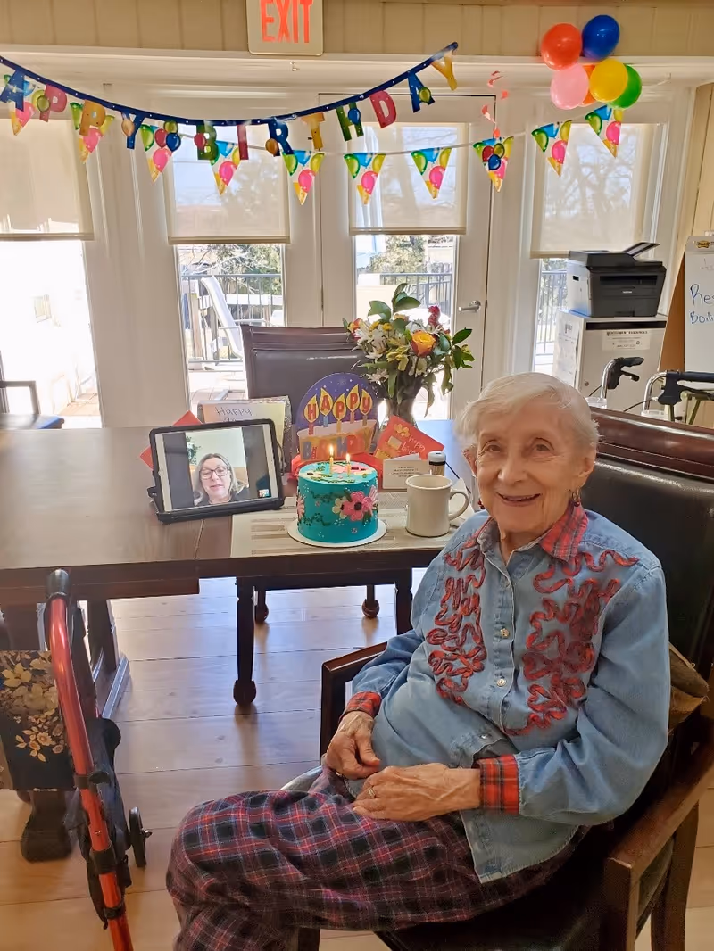 An elderly woman sitting in a chair smiling in a decorated room with a birthday banner and balloons. On the table in front of her is a birthday cake with lit candles, a bouquet of flowers, greeting cards, a coffee mug, and a tablet showing a video call with a woman. A walker is positioned beside her chair.