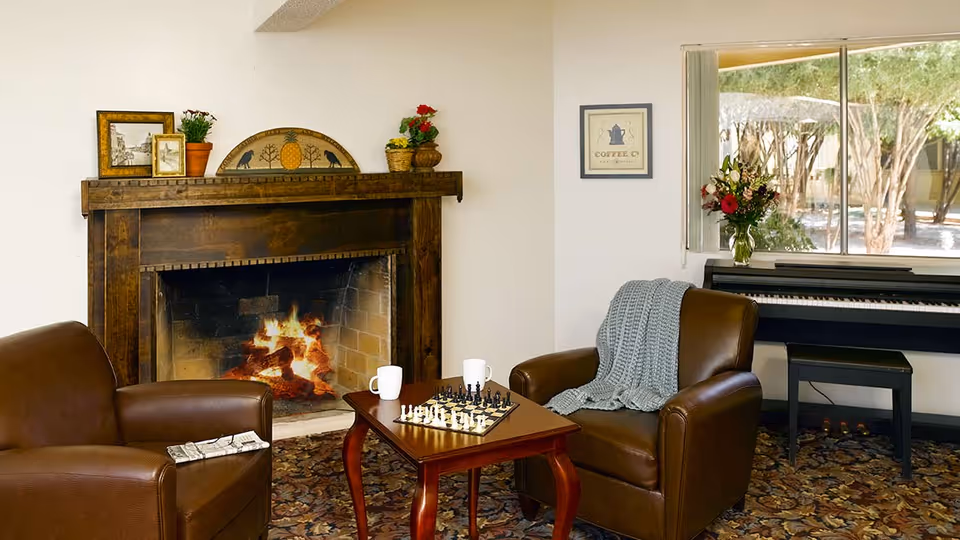 Cozy living room area with two brown leather armchairs facing a wooden coffee table with a chess set and two white mugs. A lit fireplace with a wooden mantle decorated with framed pictures and small plants is in the background. To the right, there is a piano with a vase of flowers on top and a window showing trees outside.